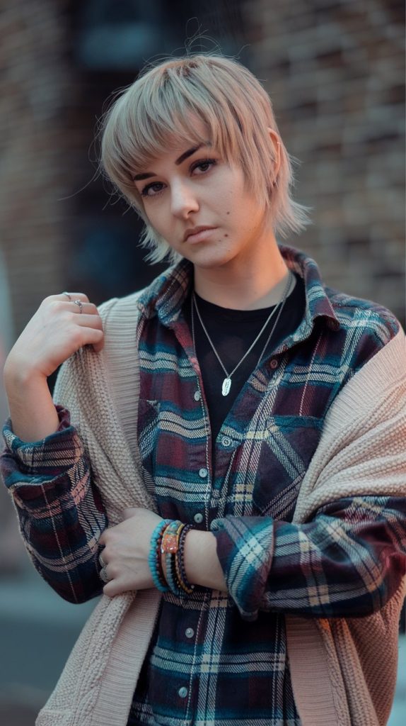 Short mullet haircut on edgy young woman, front micro bangs, longer back layers, street style outfit, colorful lighting